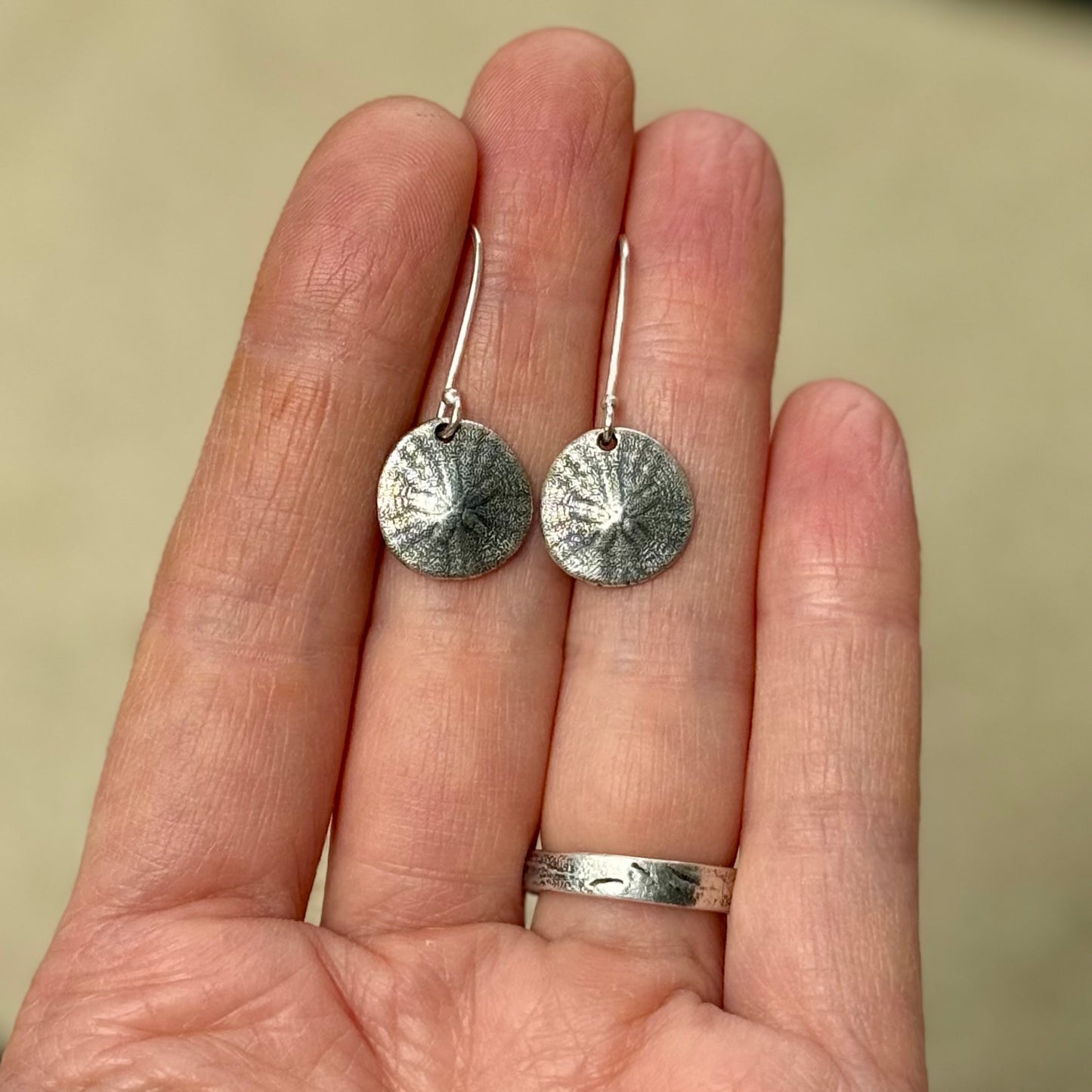 Silver sand dollar earrings held between fingers against a neutral background