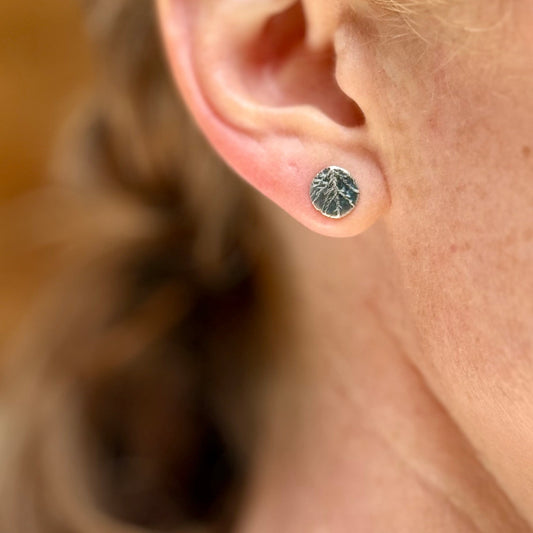 Close-up of an ear wearing a silver stud earring with a moss impression with blurred hair in the background