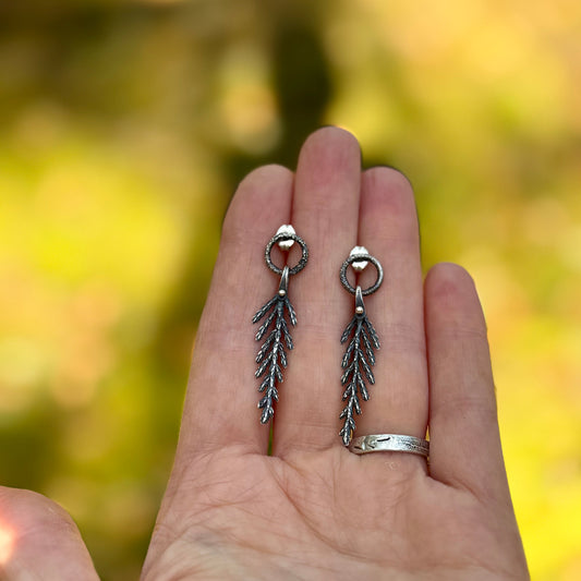 Silver cedar leaf-shaped earrings held in a hand with a blurred natural background